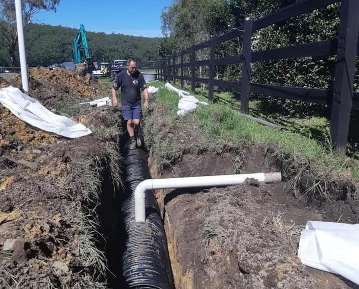 A freshly dug absorption trench with corrugated pipe and a white riser, excavator working at the far end of a fenced rural block