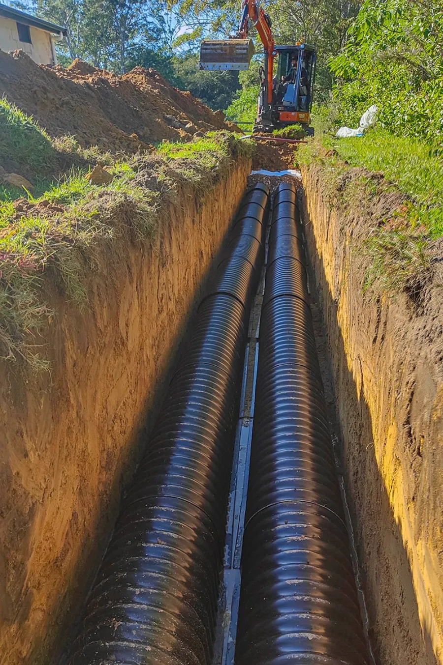 Twin corrugated drain pipes laid in a deep, freshly dug absorption trench, excavator working at the head of the run