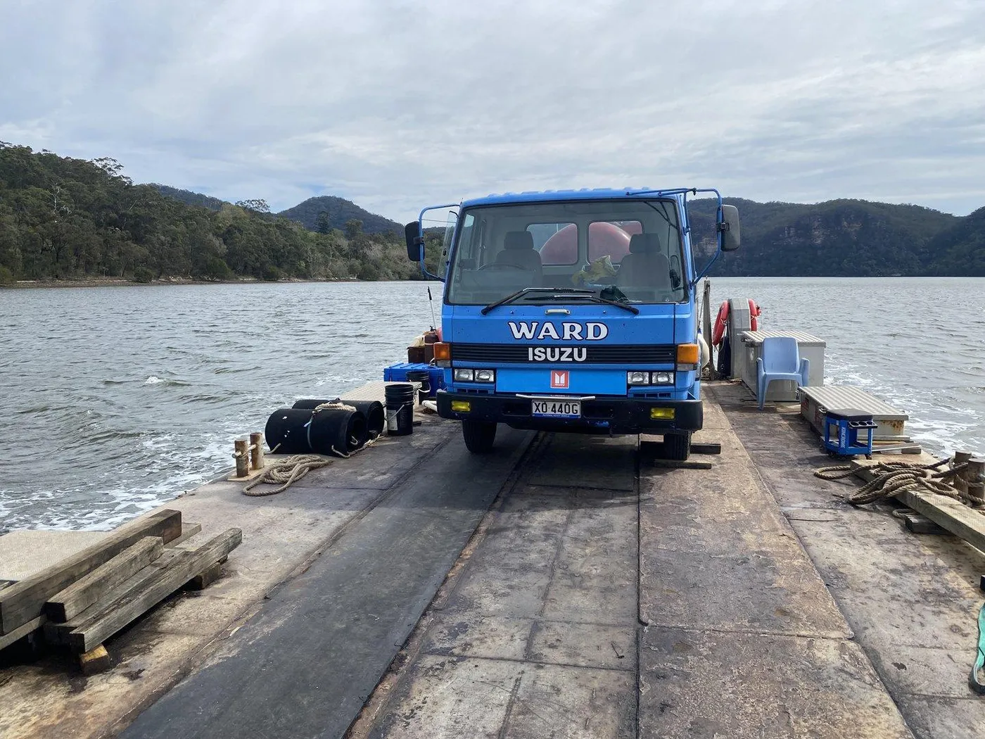 The blue Ward truck aboard a barge in the middle of the Hawkesbury River, bush ridges on both banks