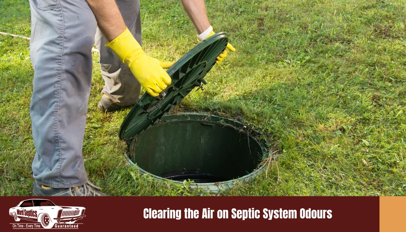 A worker wearing yellow rubber gloves lifting a heavy green plastic lid off an in-ground septic tank riser surrounded by grass.