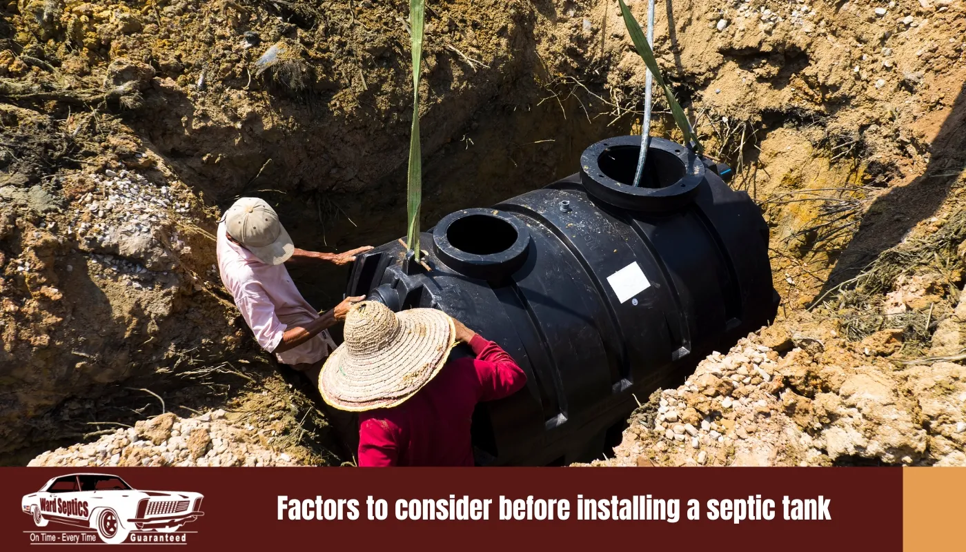 Two workers in a dirt pit positioning a large black plastic septic tank being lowered by straps