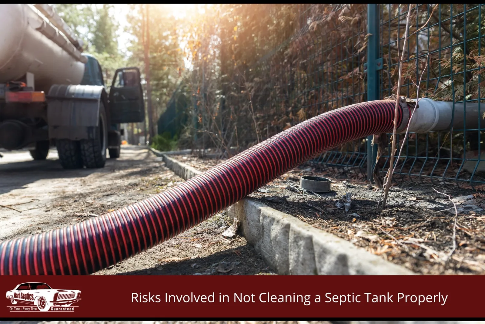 A thick, red and black corrugated vacuum hose stretching from a vacuum tanker truck parked on a driveway through a wire fence into a property.