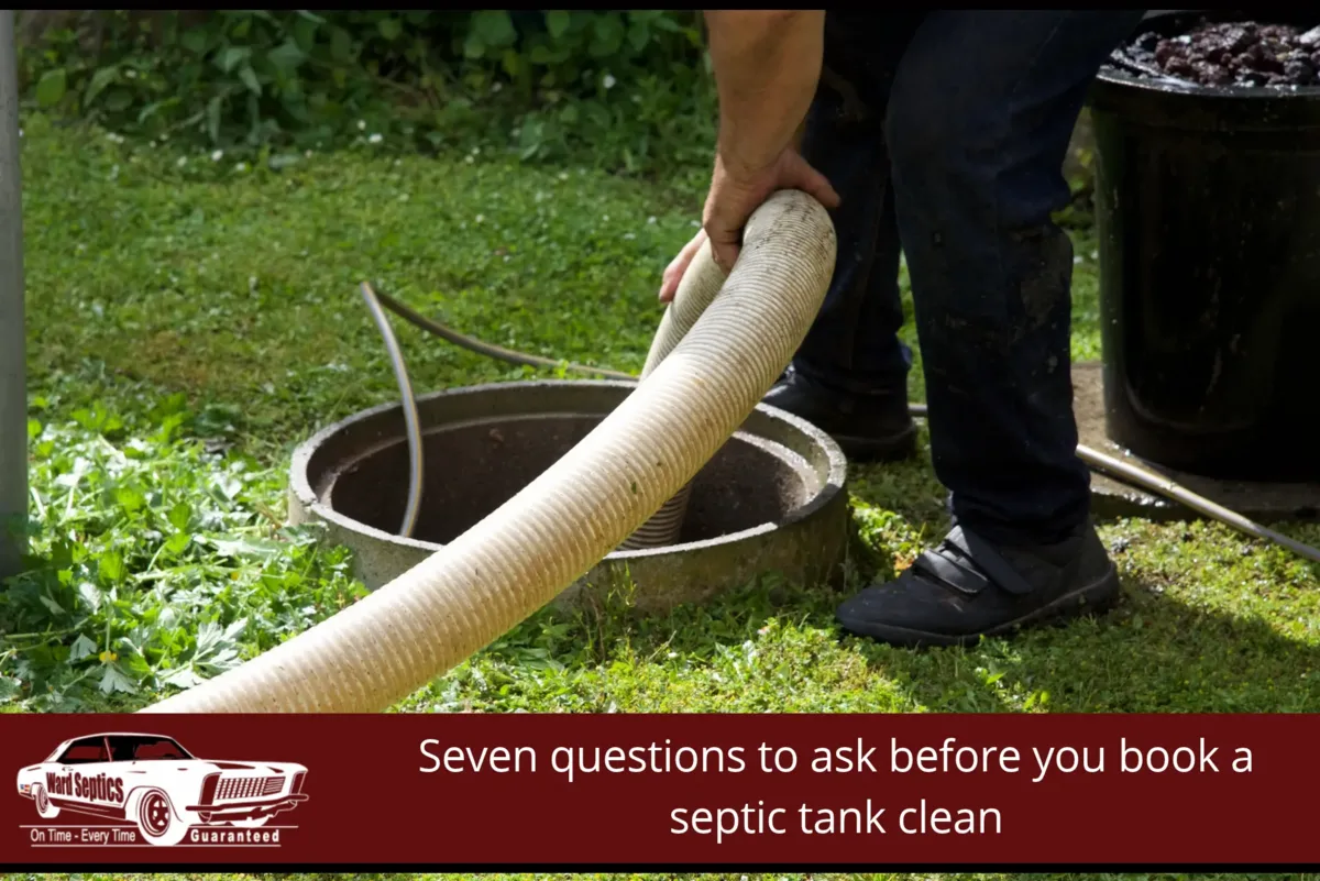 A worker in dark jeans and black shoes feeding a ribbed suction hose into an open round concrete septic tank access set in a grassy yard, beside a black drum, with the post title 'Seven questions to ask before you book a septic tank clean' and the Ward Septics logo in a red banner along the bottom