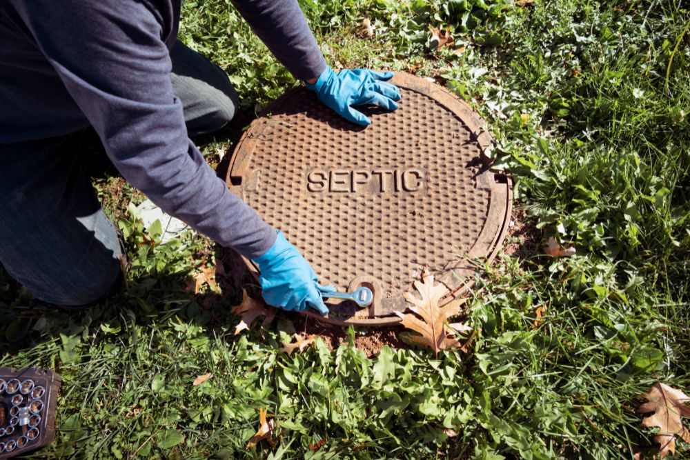 A worker wearing blue latex gloves uses a socket wrench to secure or loosen a heavy metal septic tank cover labelled "SEPTIC" in a grassy lawn.