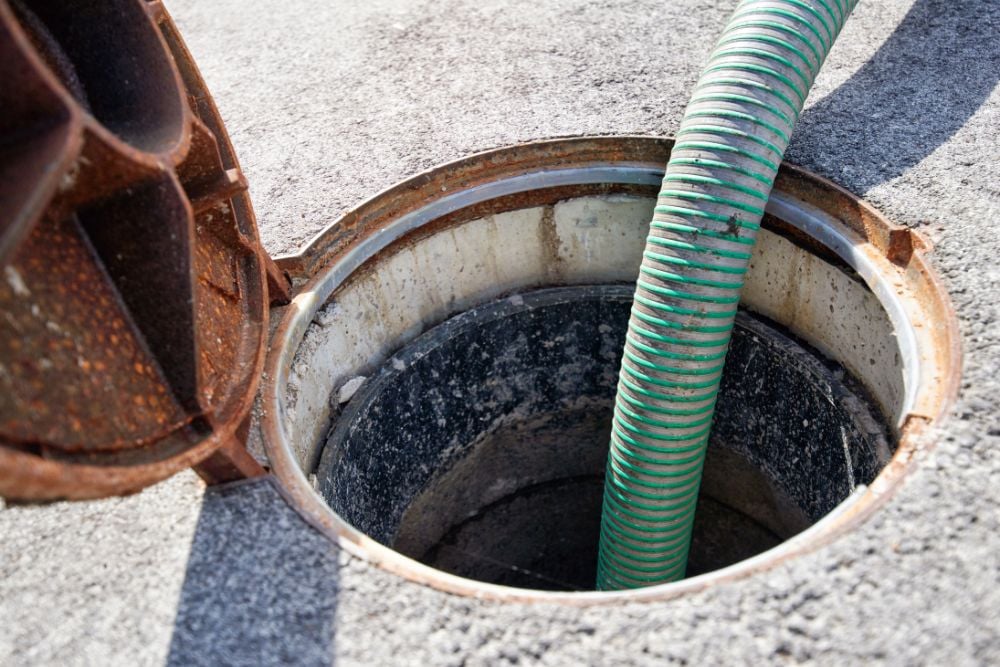 A green ribbed suction hose inserted into an open, circular manhole on an asphalt surface