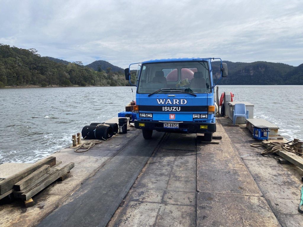 A blue Isuzu truck with "WARD" branding parked on a flat barge or ferry traveling across a river