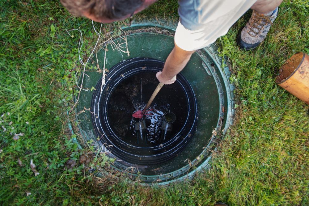 A top-down view of a worker using a long wooden tool to inspect or clear debris within an open green septic tank riser